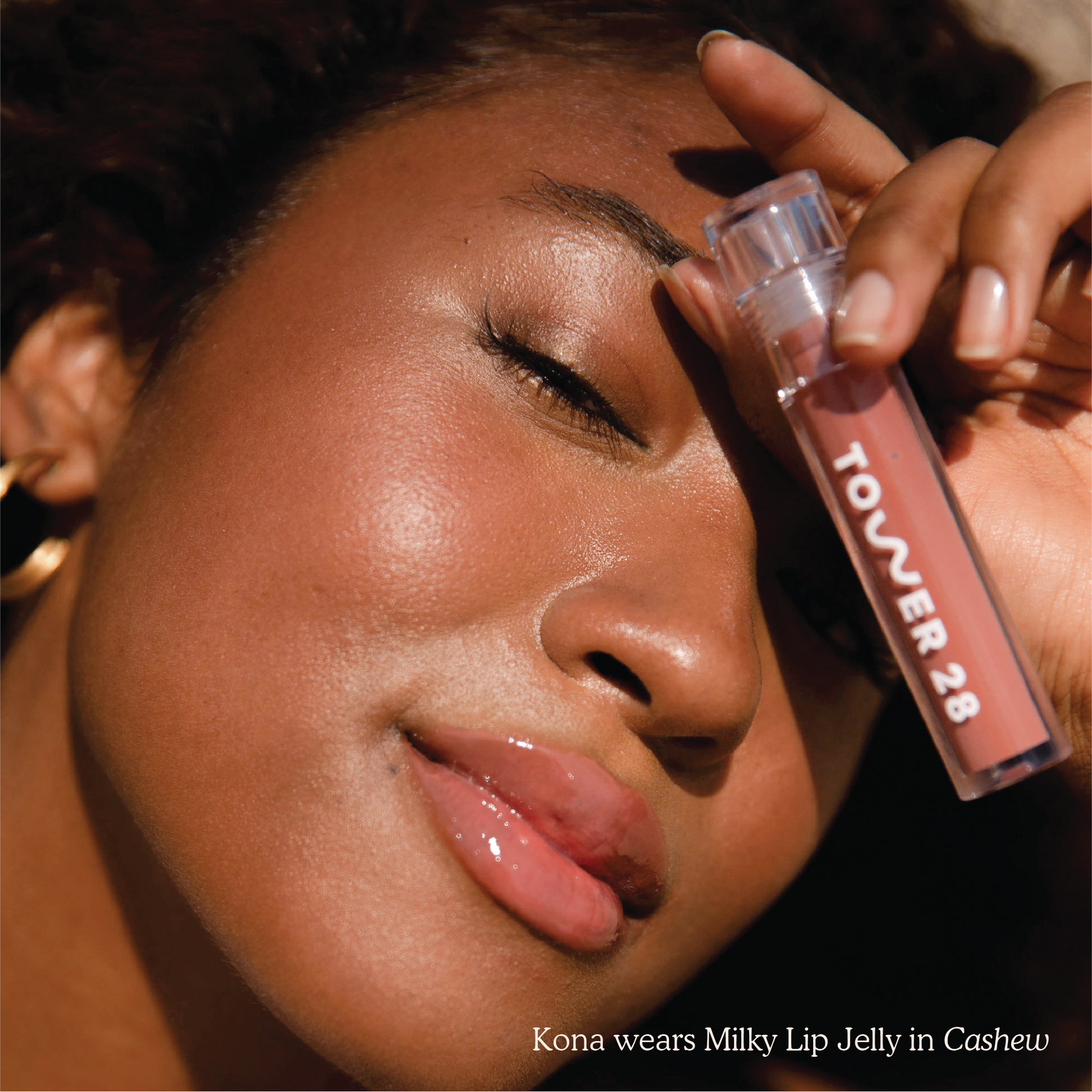 Closeup photo of a girl smiling with very glossy lips, wearing the Tower 28 Beauty ShineOn Milky Lip Jelly shade in Cashew (a milky rosy-brown), and holding an acrylic slim lip gloss tube with "Tower 28" logo over her eye