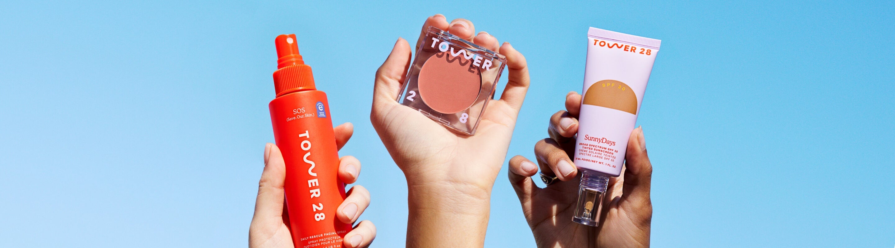 [Three hands holding up different Tower 28 Beauty products: SOS Rescue Spray, BeachPlease Cream Blush, and SunnyDays SPF 30 Tinted Sunscreen.]