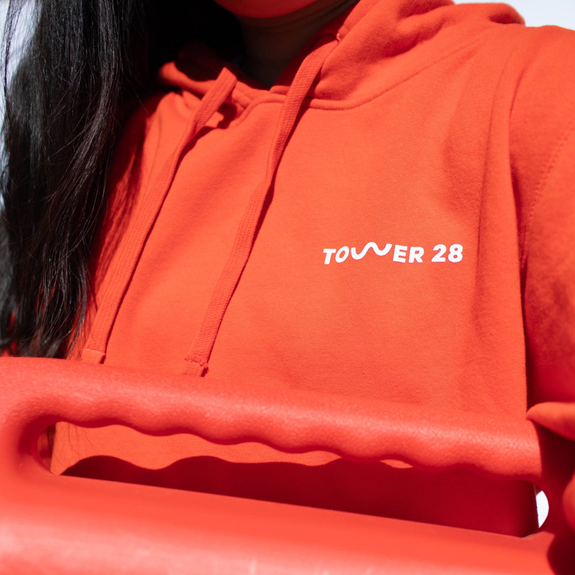 Shared: A model wearing Tower 28 Beauty's SOS Hoodie in sunset orange where all proceeds are donated to Heal The Bay.