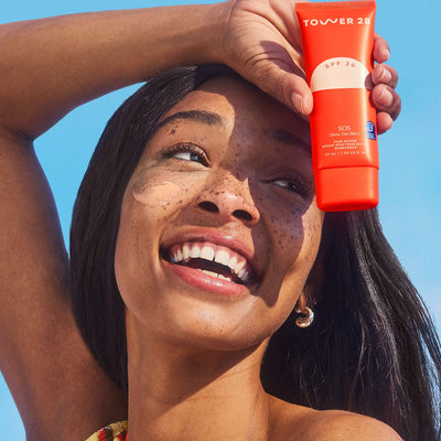 A person smiles while holding up a bottle of Tower 28 Beauty's SOS FaceGuard SPF 30. A swatch of the product is visible on their cheek.  [Shared: SOS FaceGuard SPF 30] 