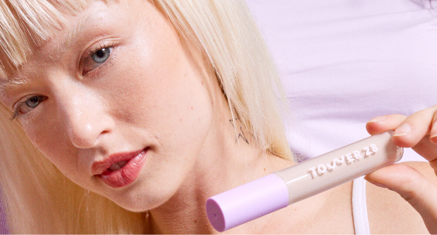 A model with blonde hair is holding a Tower 28 Beauty Swipe Serum Concealer in her hand.