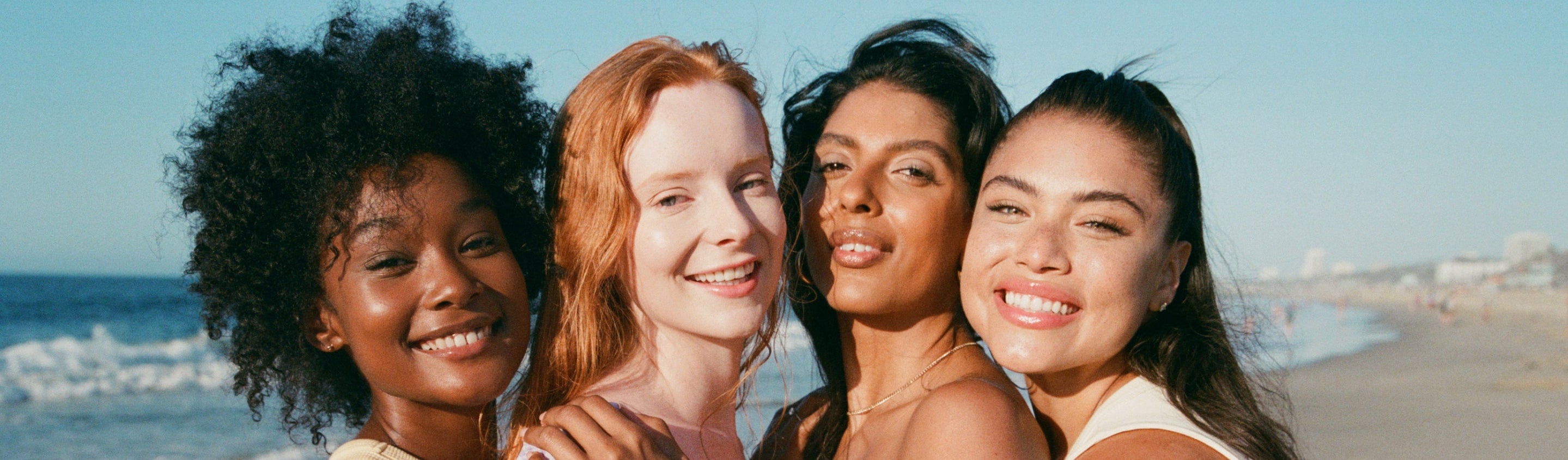 [Four models on the beach smiling at the camera wearing Tower 28 Beauty products on their faces.]