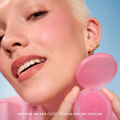 A person holds Tower 28 Beauty’s GetSet Powder Blush in the shade Malibu Cooler with a swatch of cool baby pink on their cheek. [Malibu Cooler]