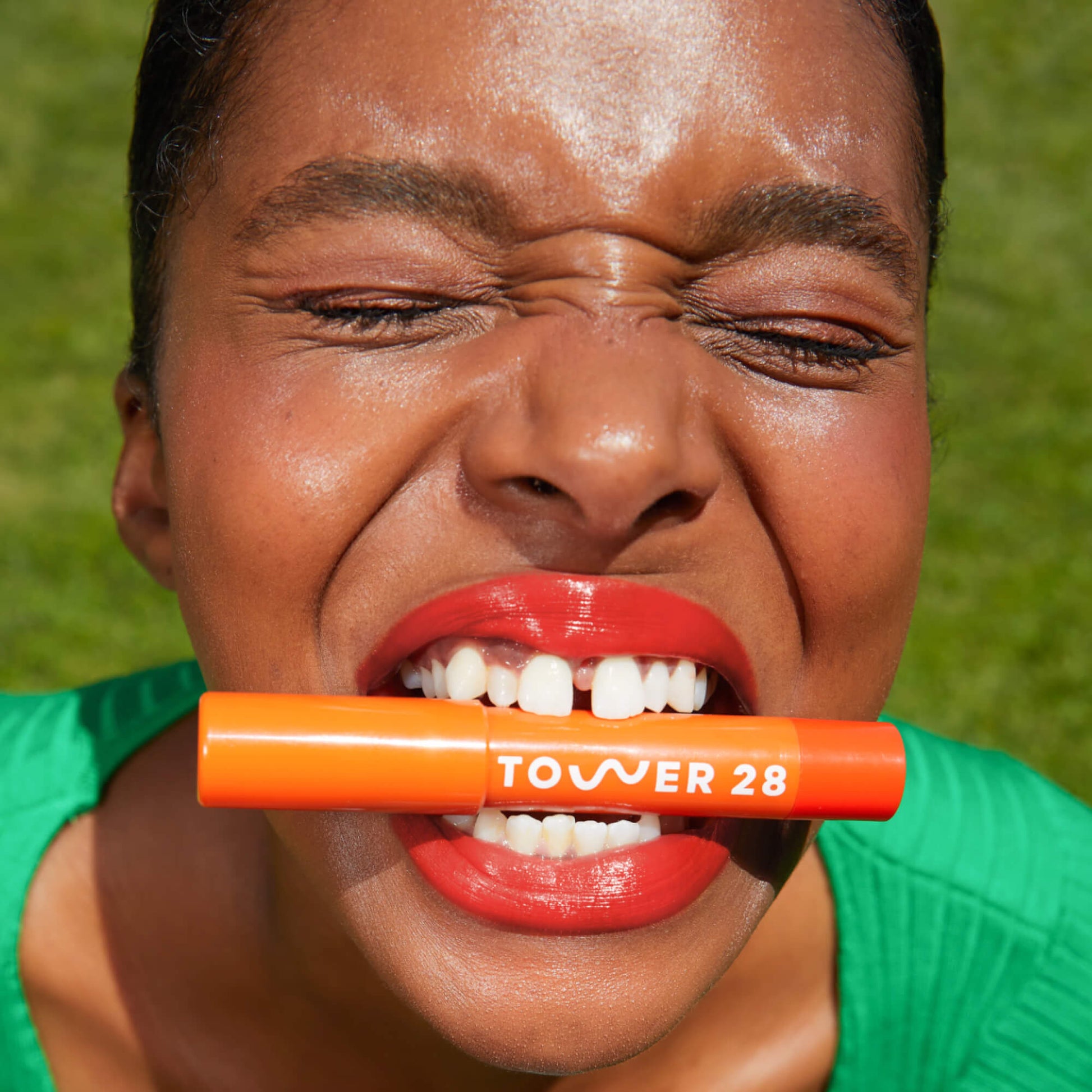 A model who has applied Tower 28 Beauty's JuiceBalm Lip Balm in shade Squeeze on her lips and is also posing with the product between her lips.