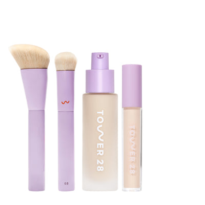 [shared] Tower 28 makeup set featuring a lavender brush, a lavender makeup brush with cream bristles, a frosted bottle of liquid foundation, and a clear tube of liquid foundation with lavender cap, all on a white background.