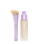 [shared] Tower 28 liquid foundation bottle in a frosted beige finish with a lavender pump, shown next to a matching lavender makeup brush with beige bristles on a white background.