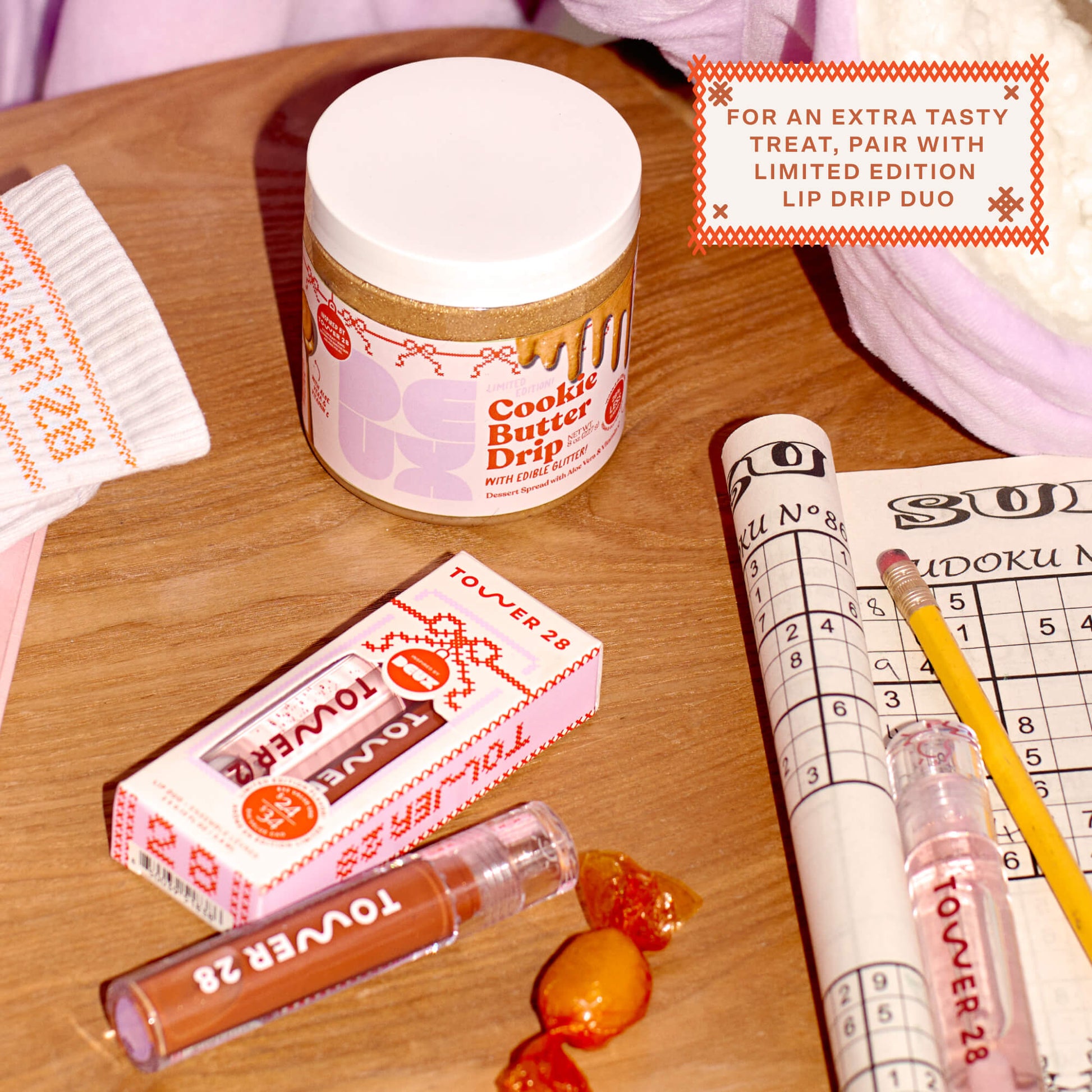 Shared: stylized flat lay of Tower 28 Beauty's 2023 limited edition Holiday Drip Collection, including Tower 28 Lip Drip Lip Gloss Duo, Tower 28 x Deux Cookie Butter Drip, and Tower 28 Toasty Toes Set Active Socks.
