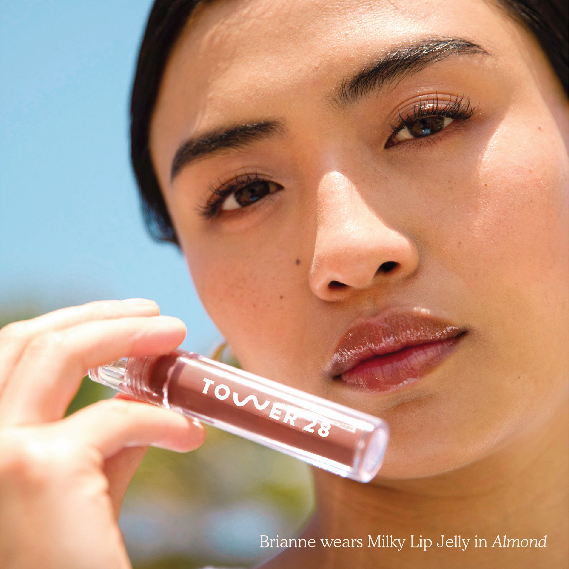 Closeup photo of a girl holding a Tower 28 Beauty ShineOn Milky Lip Jelly in shade Almond (a milky chocolate brown) in an acrylic slim lip gloss tube with "Tower 28" logo over her her chin.