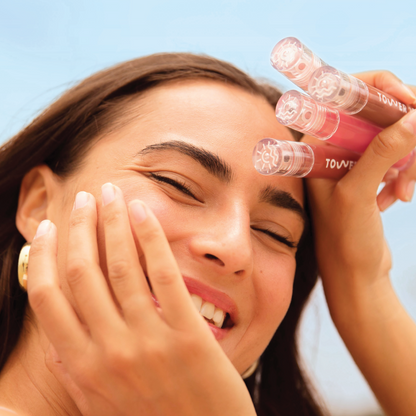 [Shared - Closeup photo of a girl with brown hair smiling, holding up 4 Tower 28 Beauty ShineOn Milky Lip Jellies (assorted shades) to her face]